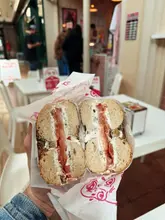 A close-up, first-person shot of a bagel held in hand, showcasing the cream cheese and tomato filling inside a branded wrapper, captured within a busy indoor market environment.