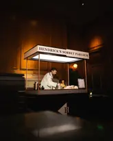 A high-quality, atmospheric photograph capturing a branded 'Hendrick's Sorbet Parlour' cart positioned within a dimly lit, wood-paneled venue. A bartender is visible behind the cart, which is set up for an exclusive event, creating a sophisticated and inviting mood.