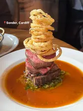 A vertical, close-up shot of a tall, stacked steak dish featuring fillet of sirloin tournedos topped with onion rings, served in a pool of sauce.