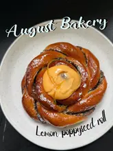 A top-down, centered photograph of a lemon poppyseed roll on a simple plate, featuring text overlay identifying the venue and the pastry.
