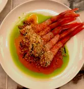 A high-angle, close-up shot of a fine-dining prawn dish featuring vibrant red prawns served over a split green and red sauce, garnished with a textured crumb topping.