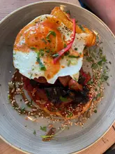 A top-down, overhead shot of a brunch dish featuring a fried egg, black pudding (morcilla), and a red sauce base, served on a rustic plate on a wooden table.
