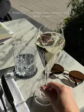 A close-up shot of a glass of sparkling wine held in a hand on an outdoor marble table, accompanied by sunglasses and a glass of water, capturing a sunny, relaxed atmosphere.