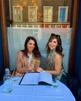 A photo of two women toasting with champagne at a restaurant table with a white tablecloth, featuring an open menu. The setting appears to be a classic French bistro or restaurant.