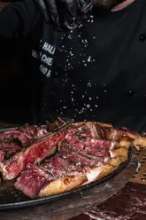 A high-quality, dramatic food photography shot featuring a chef seasoning a sliced T-bone steak. The lighting is moody and focused, highlighting the texture and juiciness of the meat, while the action of the salt falling adds a dynamic element to the composition.