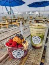 A casual, outdoor shot of an acai bowl and a matcha latte on a wooden picnic table at a cafe, with a view of a harbour in the background.