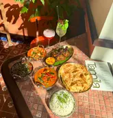 An overhead flat-lay shot of a spread of South Indian and Sri Lankan dishes, including curries, rice, roti, and cocktails, set on a tiled table in a sunlit restaurant.