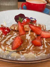 A close-up, high-quality shot of Biscoff, strawberry, and white chocolate pancakes served at a cafe. The image effectively captures the texture of the sauces and the freshness of the fruit, with a coffee cup visible in the soft-focus background.