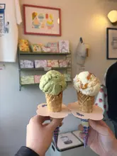 A first-person perspective shot of two ice cream cones held up in front of a shop interior, showcasing a matcha and white chocolate flavour alongside a ramen flavour.