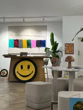 A clear, eye-level shot of the interior of Helm Gallery in Brighton, showcasing a wooden desk with a large smiley face logo, a piece of art on the wall, and cafe seating area.