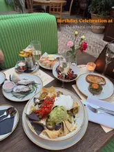 A flat-lay photograph of a table spread at a restaurant, featuring various dishes including nachos, oysters, steak tartare, and chicken wings, with text overlay indicating a birthday celebration.