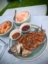 A top-down, well-composed shot of a homemade Thai meal featuring a whole fried black tilapia as the centerpiece, accompanied by papaya salad, lemongrass jasmine rice, and two dipping sauces. The lighting is bright and even, effectively showcasing the textures of the crispy fish and the vibrant colors of the accompanying dishes against a neutral, clean background.