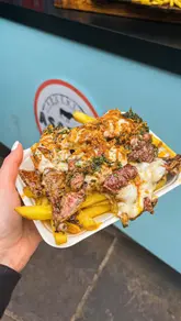 A short-form video review of Asador Argentine Grill in Camden Market, featuring the preparation and consumption of a ribeye steak sandwich and loaded fries. The creator highlights the quality of the meat, the chimichurri sauce, and the overall street food experience.