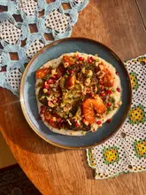 A top-down, well-lit shot of a homemade vegetarian dish featuring roasted broccoli and crispy halloumi served over a garlic and onion yogurt base, garnished with pomegranate seeds and toasted hazelnuts. The image is styled on a wooden surface with a crochet doily, evoking a cozy, home-cooked aesthetic.