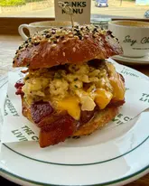 A high-quality, close-up shot of a breakfast bagel served on branded paper, likely in a cafe setting. The image effectively captures the textures of the scrambled eggs, melted cheese, and bacon, with coffee cups visible in the background to establish a relaxed breakfast atmosphere.