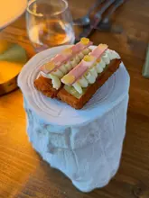 A high-quality, close-up shot of a refined appetizer served on a unique, textured white pedestal. The dish features a crispy base topped with piped cream, cured meat, and gel dots, showcasing precise, fine-dining presentation.