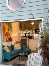 A well-composed, inviting shot of a luxury beach hut interior, framed by the open doorway. The image effectively captures the cozy, aesthetic appeal of the accommodation, using the natural light and the view of the beach to create a desirable atmosphere.