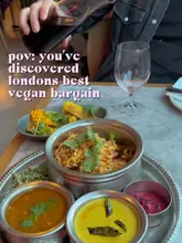 A top-down shot of a vegan thali meal at a restaurant, featuring a variety of small bowls and a main dish, with a hand pouring wine in the background.