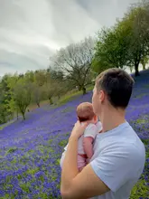 A high-quality, candid-style photograph featuring a man holding a baby, overlooking a scenic field of bluebells at Clent Hills. The image captures a peaceful, family-oriented outdoor moment, effectively showcasing the natural beauty of the location.