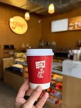 A clean, well-composed shot of a branded takeaway coffee cup held in front of a coffee shop interior. The image effectively highlights the branding while providing a sense of place through the softly blurred background.