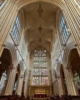 A high-quality, symmetrical architectural shot of the interior of Bath Abbey, focusing on the intricate fan-vaulted ceiling and the large stained glass East Window. The image captures the grandeur and historical significance of the Perpendicular Gothic structure, with natural light filtering through the glass to illuminate the nave.