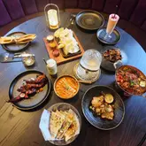 A high-quality, overhead flat-lay photograph of an extensive Indian dining spread. The image features a variety of dishes including curries, lamb chops, naan, and mocktails, arranged symmetrically on a dark, textured table. The lighting is soft and even, highlighting the textures and vibrant colours of the food without creating harsh reflections or shadows.