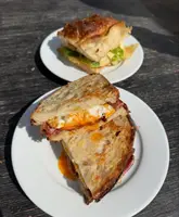 A top-down shot of two breakfast sandwiches on white plates, set against a dark wooden table. The image features a bacon and egg sandwich on sourdough in the foreground and a focaccia sandwich in the background, captured in bright, direct sunlight.