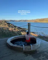 A high-quality, aspirational travel image featuring a person relaxing in an outdoor wooden hot tub on a deck, overlooking a scenic Scottish loch. The image effectively captures the serene, luxury nature of the accommodation, using natural golden hour lighting to highlight the stunning landscape and the unique amenities offered by the venue.