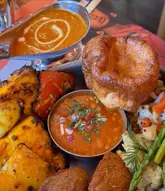 A close-up, top-down shot of an Indian-fusion Sunday roast featuring paneer tikka, a Yorkshire pudding, roasted vegetables, and a side of curry sauce.