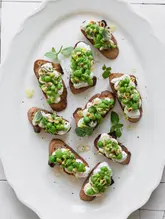 A high-quality, overhead food photography shot featuring several crostini topped with ricotta, smashed peas, pine nuts, and lemon zest on a white platter. The lighting is bright and even, highlighting the vibrant green of the peas and the creamy texture of the ricotta, creating a fresh and inviting aesthetic suitable for a Mediterranean-inspired menu.