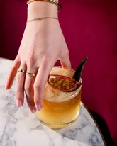 A professionally shot, high-end image of a cocktail being held by a hand, featuring a passion fruit garnish and a marble surface against a deep red backdrop.
