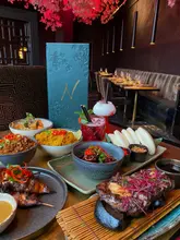 A high-quality, atmospheric promotional shot of a table spread at Noshi, featuring a variety of Japanese and Chinese-inspired dishes including steak, noodles, bao buns, and cocktails, set against a moody, cherry-blossom-themed restaurant interior.