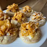 A top-down, slightly angled shot of a batch of homemade cornflake tart cookies on a white plate, set against a wooden background. The cookies are topped with a cornflake mixture and jam, presenting a rustic, home-baked aesthetic.
