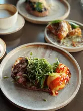 A high-quality, well-lit food photography shot featuring a brunch spread, with the primary focus on a steak and eggs dish on sourdough, accompanied by other brunch items in the background.