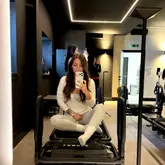 A mirror selfie taken inside a Lagree/Pilates studio, featuring the creator sitting on a reformer machine. The image captures the aesthetic of the studio with its dark, modern interior and LED lighting, documenting a fitness experience.