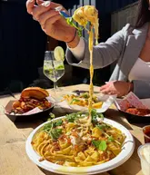 A close-up, eye-level shot of a person lifting a forkful of pasta, with a table spread of street food including a burger, fries, and a cocktail in the background. The scene is set outdoors in bright, direct sunlight.