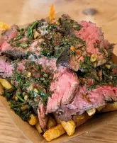 A close-up, top-down shot of a street food dish consisting of sliced steak with chimichurri sauce served over a bed of fries in a cardboard tray.