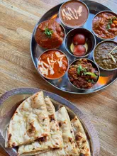 A top-down, overhead shot of an Indian thali set featuring multiple small bowls of various curries and a side of naan bread on a wooden table. The lighting is natural and even, highlighting the textures and colours of the food.