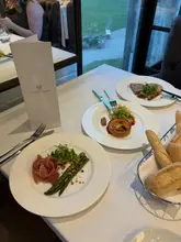 A high-angle shot of a table setting at Cameron House, featuring three plates of food including asparagus with prosciutto, a tartlet, and a terrine, alongside a menu card and a basket of bread.