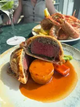 A close-up, high-angle shot of a Beef Wellington served with a fondant potato and sauce, with a plate of langoustines and a cocktail visible in the background at a restaurant.