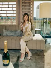 A polished lifestyle shot featuring a woman in luxury pajamas holding a glass of champagne in a hotel suite, with a bottle of Perrier-Jouët in the foreground.