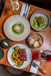 A high-quality, professional flat-lay photograph of a fine dining dinner spread, featuring a rump steak, asparagus velouté, stone bass, and a bread basket, set on a rustic wooden table.
