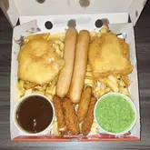 Here are a few options for your SEO alt text (all under 125 characters):

**Option 1 (Focus on keywords):**
A Munchie Box takeaway featuring fish fillets, sausages, chicken strips, chips, gravy, and peas from Nick’s Wok in Chester.

**Option 2 (Descriptive):**
A large takeaway Munchie Box from Nick’s Wok and Fish Bar in Chester filled with golden chips, fish, sausages and chicken.

**Option 3 (Concise):**
Close up of a sharing Munchie Box containing fish, chips, sausages, and chicken strips from Nick’s Wok in Chester.