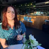 A portrait of a woman holding a glass of white wine at a rooftop bar during golden hour, with the venue's neon sign visible in the background.