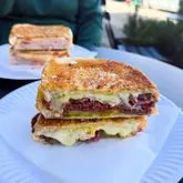 A high-quality, close-up shot of two toasted sandwiches served on paper plates, likely in an outdoor or casual market setting. The focus is sharp on the cross-section of the front sandwich, effectively highlighting the melted cheese and fillings, making the food look very appetizing.