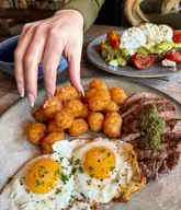 A high-quality, overhead shot of a brunch spread featuring steak and eggs with crispy tater tots in the foreground, and avocado toast with poached eggs in the background. The image uses natural lighting and a shallow depth of field to highlight the textures of the food, with a hand reaching into the frame to add a sense of action and engagement.