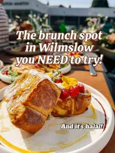 A promotional image for a brunch venue featuring a plate of French toast topped with berries and edible flowers. The image uses natural light and a shallow depth of field to highlight the food, though the composition is slightly cluttered by the surrounding table setting.