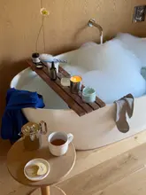 A lifestyle shot capturing a relaxing morning routine in a hotel or holiday stay, featuring a bathtub with bubbles, a wooden bath tray with skincare products, and a side table with tea and a biscuit.