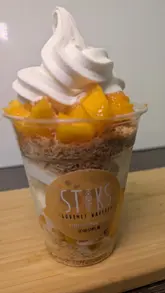 A close-up, satisfying video showing the assembly of a mango-based dessert in a cup, featuring layers of shredded pastry, soft-serve ice cream, and syrup.
