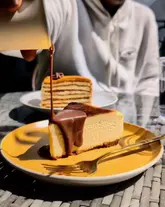 A close-up, high-angle shot capturing a decadent chocolate ganache pour over a slice of Basque cheesecake. The image is well-lit with natural light, highlighting the texture of the cheesecake and the glossy finish of the chocolate sauce. The background features a blurred figure, suggesting a casual dining environment.